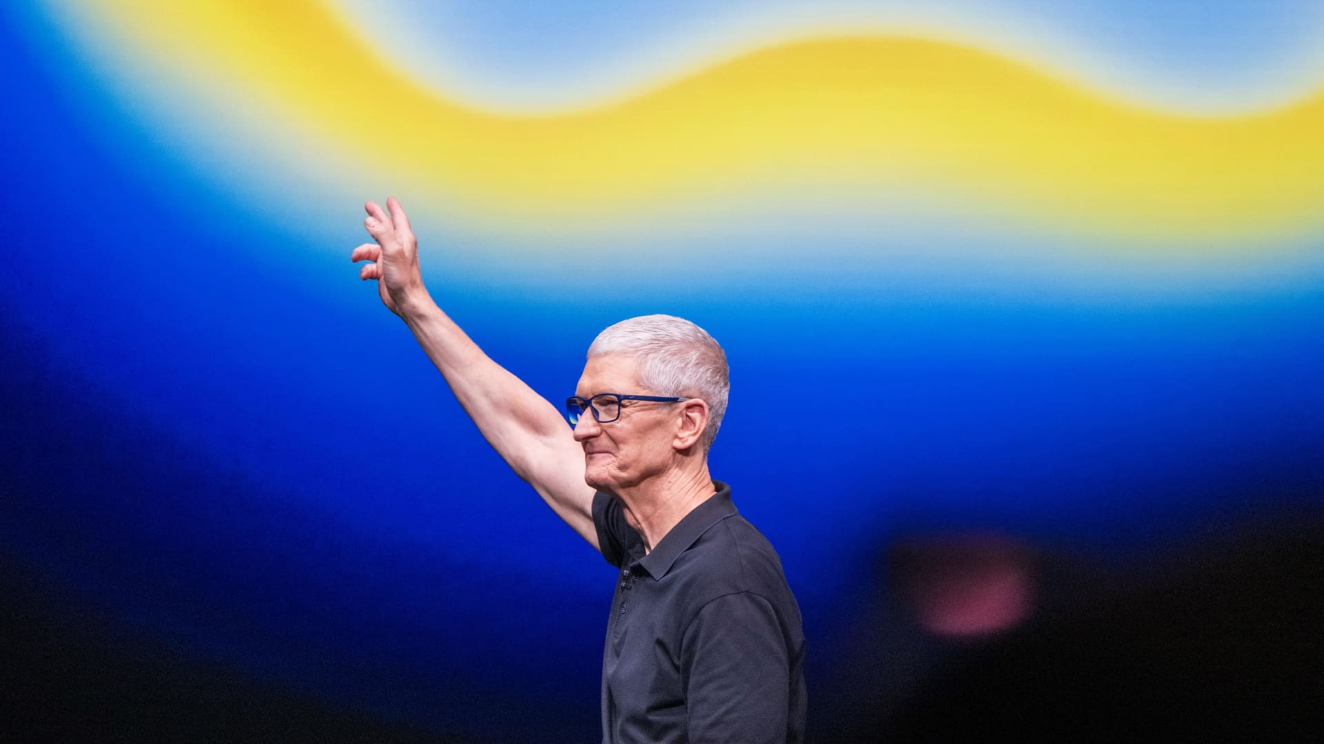Tim Cook, cheveux argentés et lunettes, salue le public sur la scène de la WWDC 2026 devant un fond bleu et jaune.